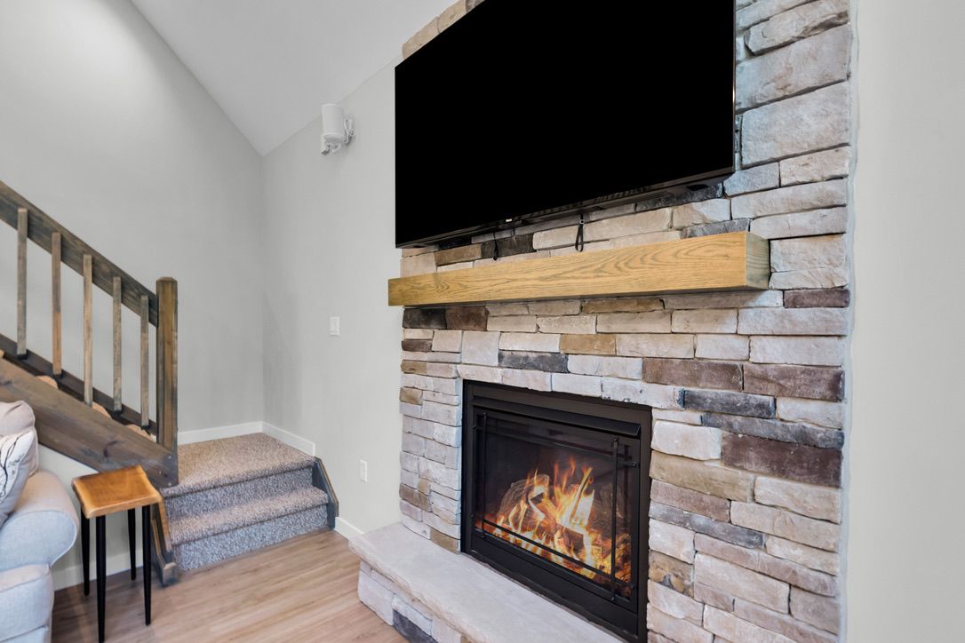 A modern living room with a stone fireplace, a flat-screen TV mounted above it, and a staircase leading to another level.