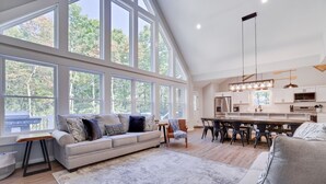 A spacious living room with a large window, a sofa, a coffee table, and a dining area with chairs and a table. - Lake & Beach + Firepit + King Beds + Swing set (Jim Thorpe)