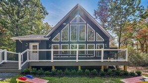 A dark-colored cabin with a balcony, surrounded by trees and a grassy area. - Lake & Beach + Firepit + King Beds + Swing set (Jim Thorpe)
