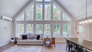 A spacious room with a high ceiling, large windows, a wooden dining table, and a sofa with cushions. - Lake & Beach + Firepit + King Beds + Swing set (Jim Thorpe)