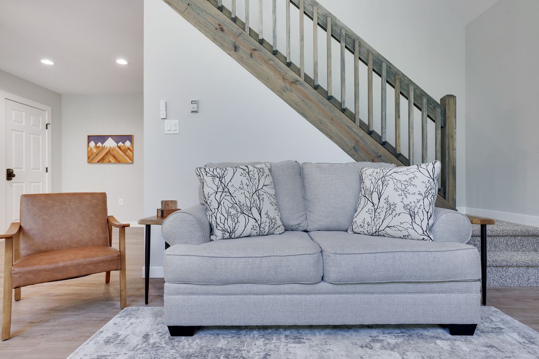 A living room with a grey sofa, a brown leather chair, a wooden staircase, and a painting on the wall.