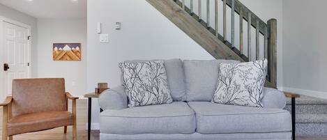 A living room with a grey sofa, a brown leather chair, a wooden staircase, and a painting on the wall.