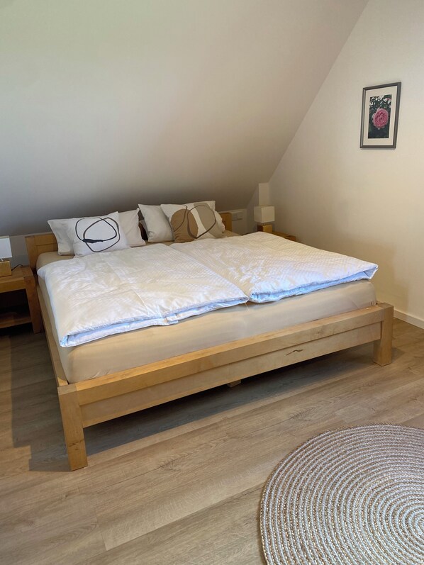 A neatly made bed with a wooden frame, a white duvet, and several pillows. There is a small bedside table with a lamp and a framed picture on the wall. - Neue , Moderne , Lichtdurchflutete Ferienwohnung 🌸 (Hallerndorf)