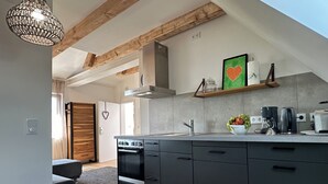 A modern kitchen with dark cabinetry, a built-in oven, and a coffee maker. There is a wooden ceiling and a hanging light fixture. - Neue , Moderne , Lichtdurchflutete Ferienwohnung 🌸 (Hallerndorf)