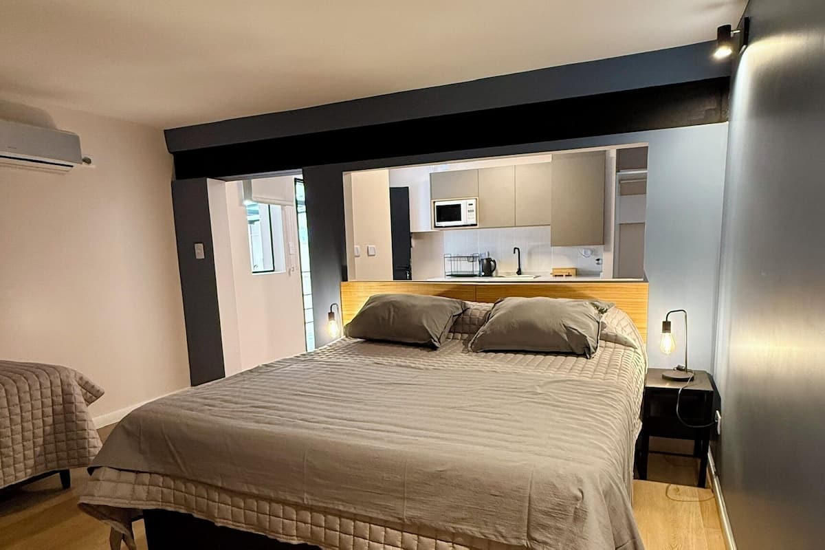 A modern bedroom with a large bed, a bedside table with a lamp, and a built-in kitchen area with a microwave.