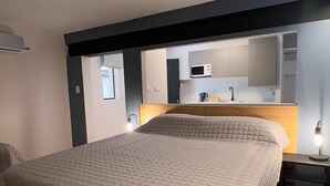 Et moderne soveværelse med en stor seng, trægulv, indbygget skrivebord og et tekøkken i baggrunden.