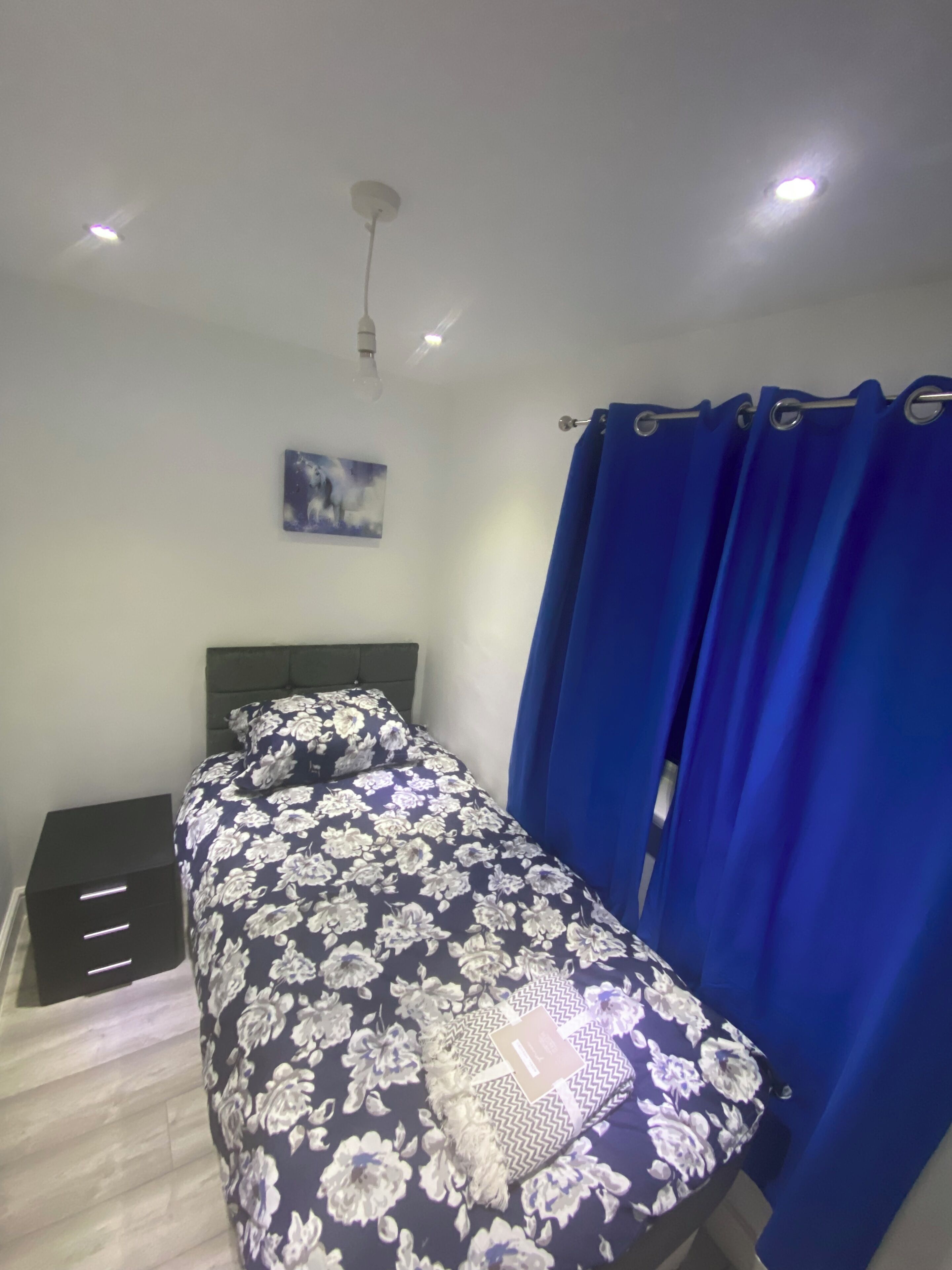 A bedroom with a bed, a nightstand, a wall-mounted light, a ceiling fan, and a window with blue curtains.