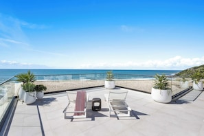 A terrace with a clear view of the beach and ocean, featuring potted plants and lounge chairs.