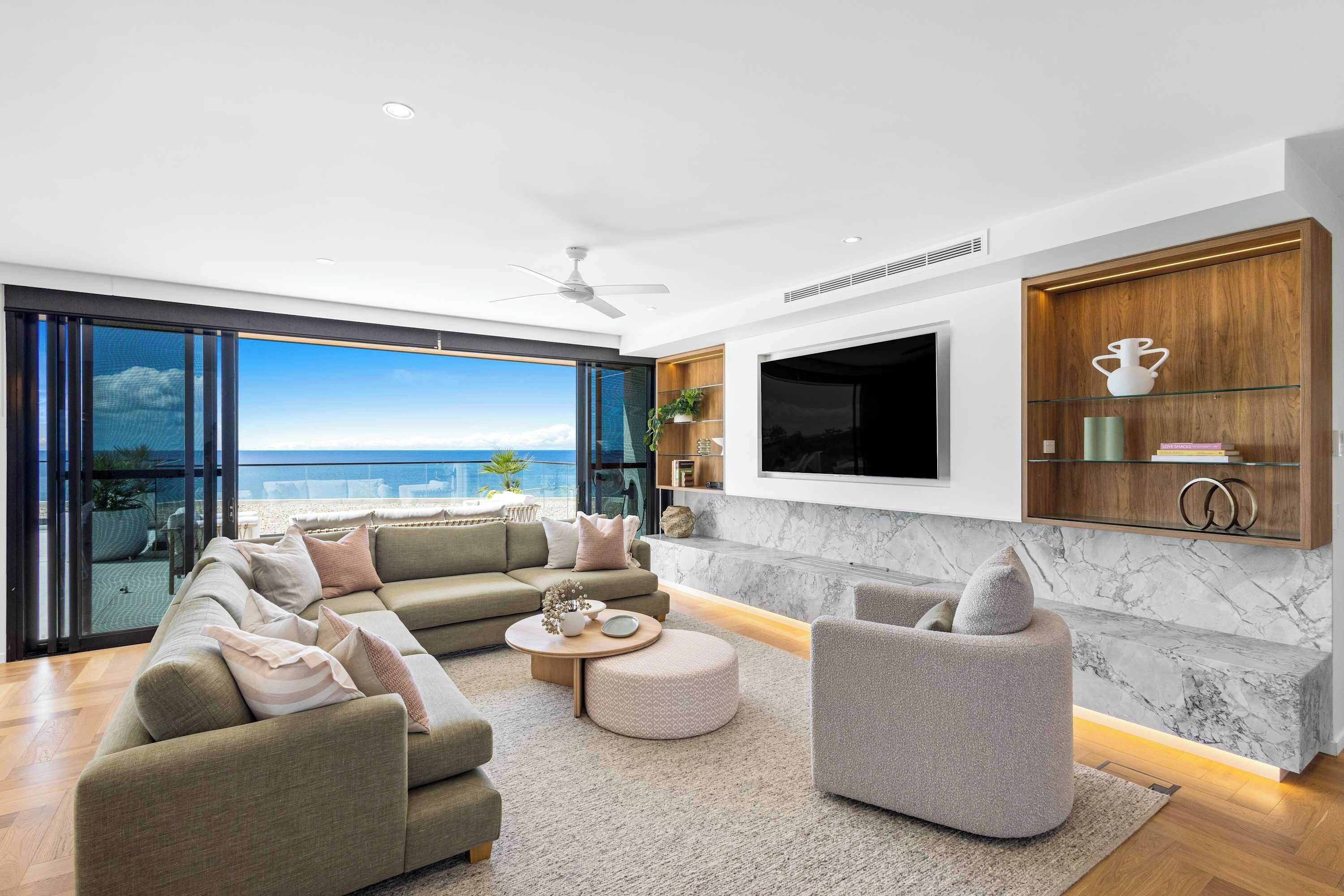 Un salon moderne doté d’une grande fenêtre donnant sur une plage, d’un téléviseur à écran plat fixé au mur et d’un meuble en bois avec des étagères en verre.