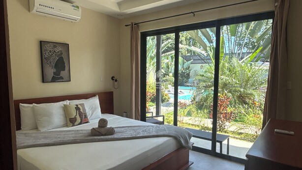 Une chambre d’hôtel avec un lit, un climatiseur, un tableau encadré et une vue sur une piscine et des espaces verts.