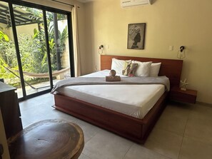 Ein Schlafzimmer mit einem Bett, einem Nachttisch, einem Holzschemel, einem Fenster mit Blick auf Grünflächen und einem Wandbild.