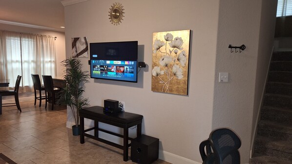 A living room with a flat-screen TV mounted on the wall, a decorative painting, a small table with a speaker, and a potted plant.