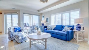 Un salón amplio con un sofá azul, sillones estampados, una mesa de centro blanca y vistas de la ciudad a través de amplias ventanas.