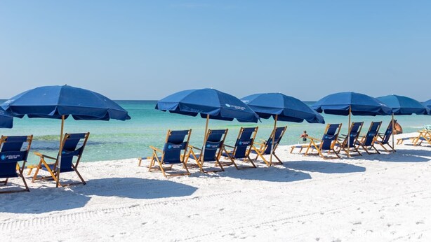 A beach with rows of blue beach chairs and umbrellas, a clear sky, and a calm sea. - The Brinley House:Steps from Beach/Pool/Bikes etc. (Destin)