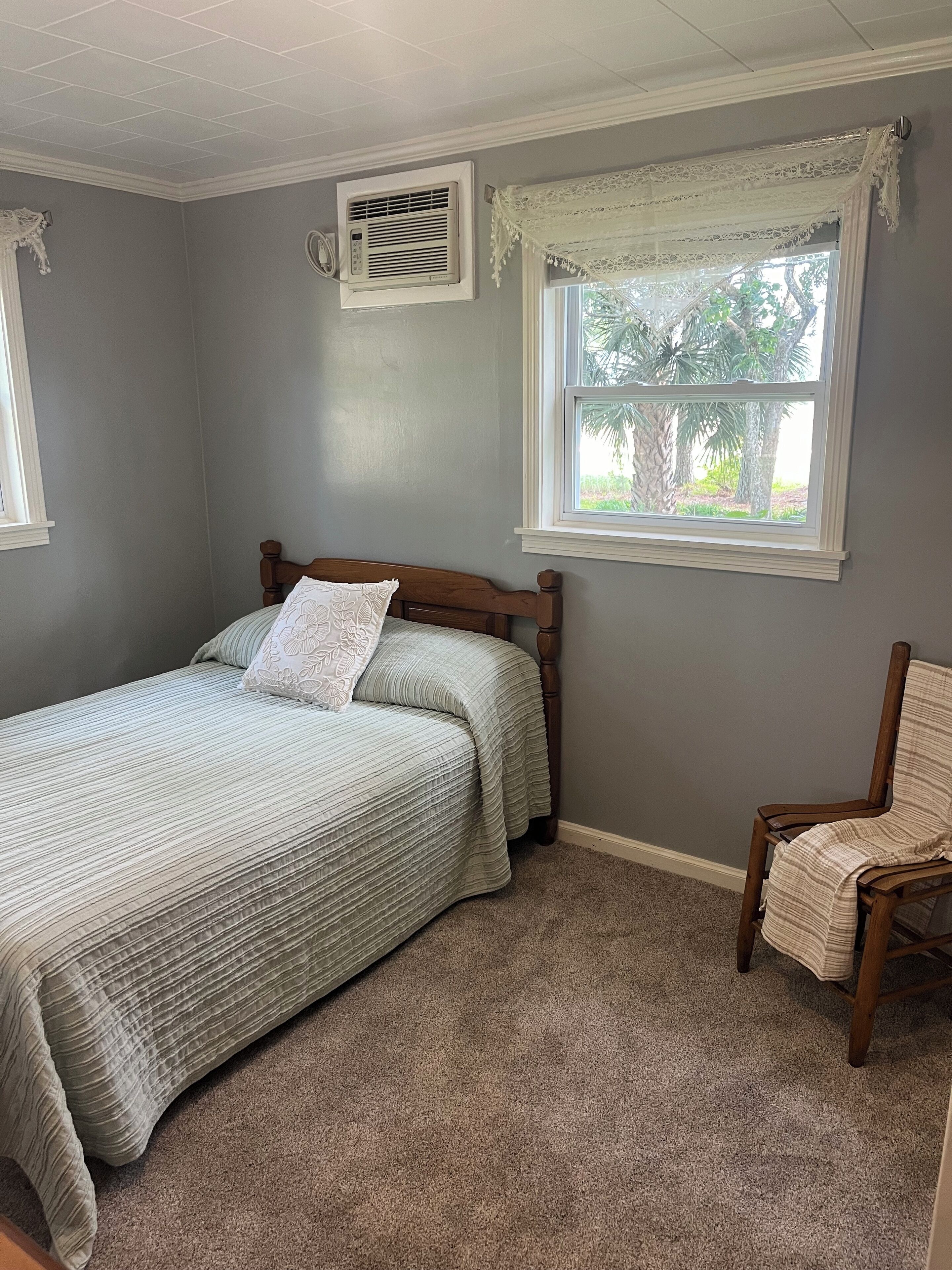 Un dormitorio con cama, una silla, una ventana con cortinas de encaje y un equipo de aire acondicionado.