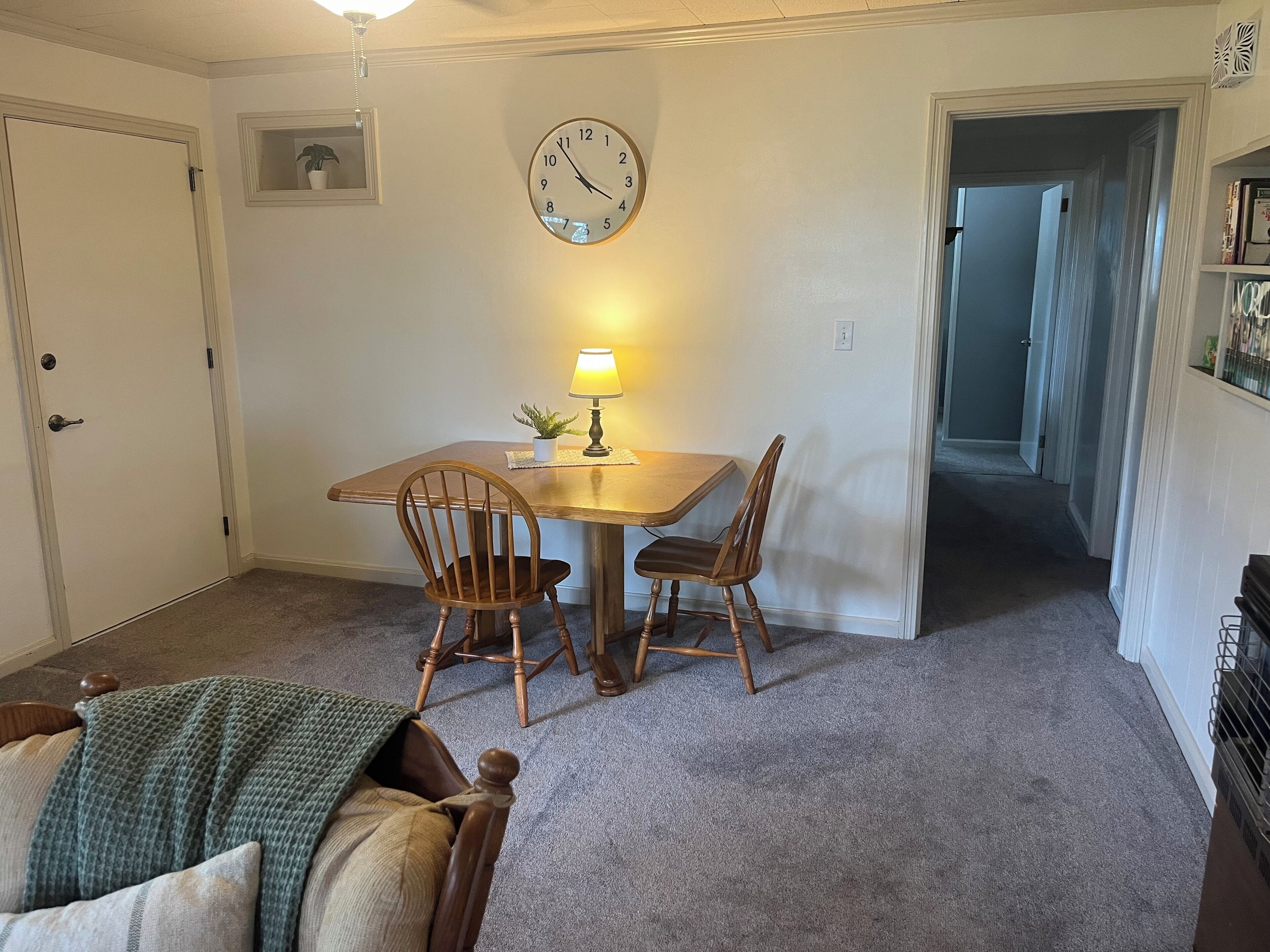 A dining area with a wooden table and two chairs, a wall clock, and a small potted plant on the table.