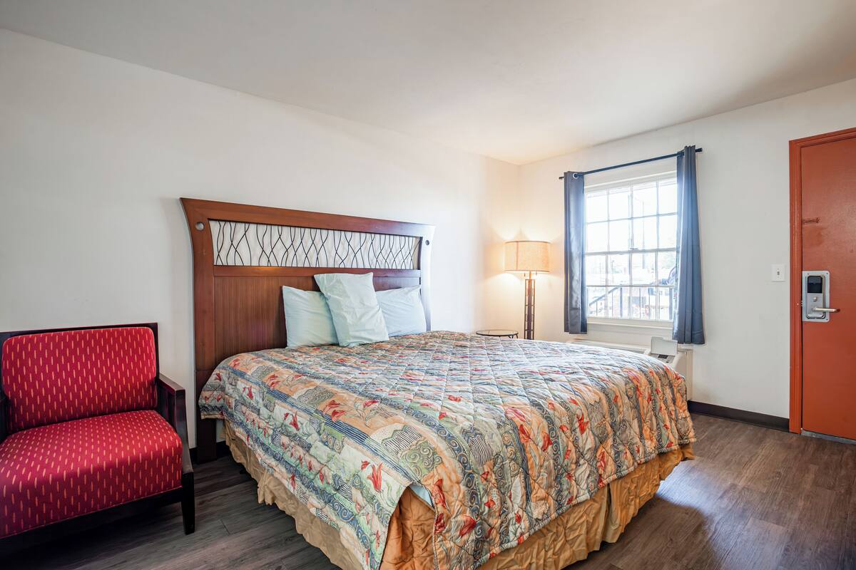 A hotel room with a bed, a red chair, a window with curtains, and a door.
