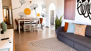 A modern living room with a gray sofa, a dining table with chairs, a large wall art piece, and a hanging wicker light fixture.