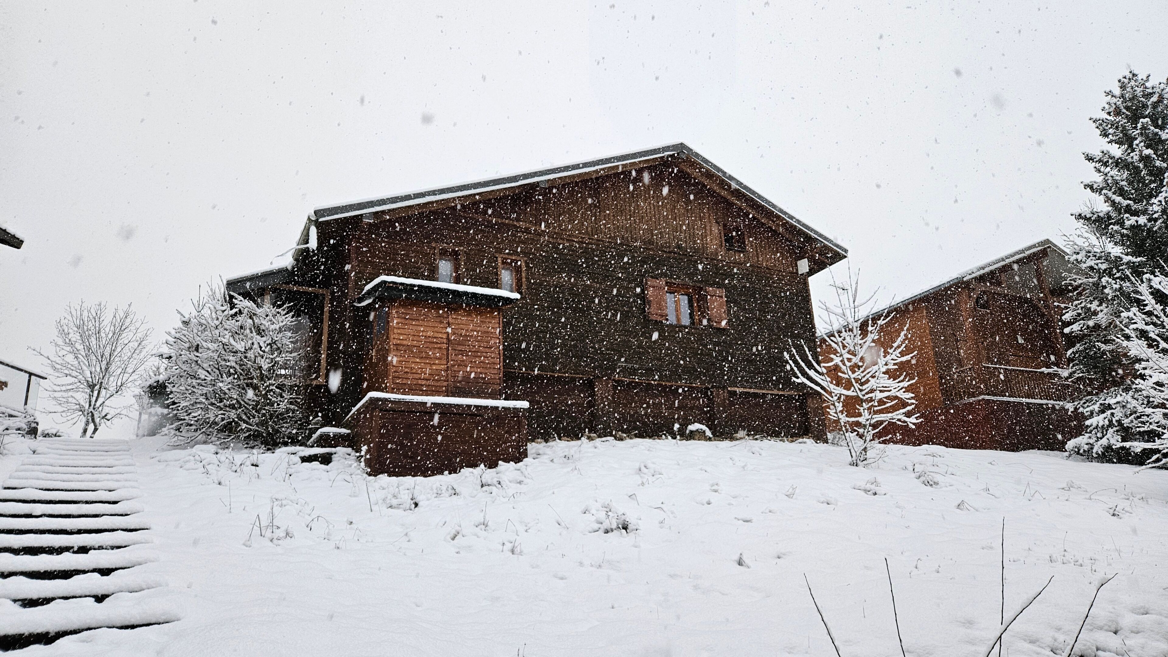 A wooden cabin with a steep roof, surrounded by snow-covered ground and trees.