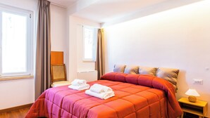 A bedroom with a large bed, wooden flooring, a nightstand with a lamp, and a window with curtains.