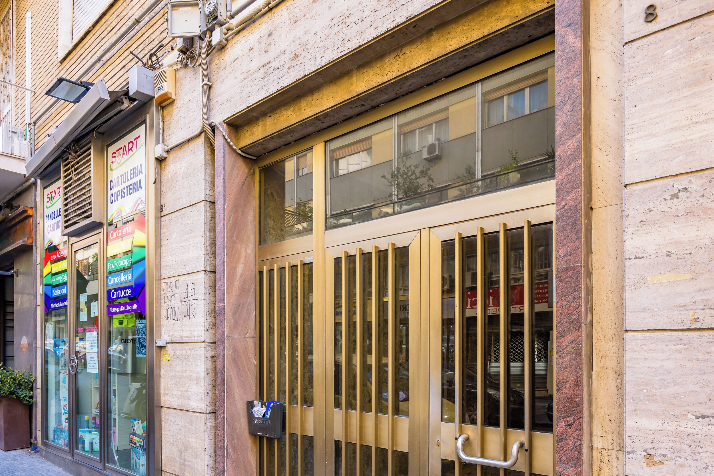 A building facade with a large, barred entrance door, glass storefronts with signage, and a visible fire hose on the wall.