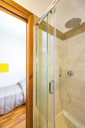 Enjoy a compact bathroom with a glass-enclosed shower, handheld showerhead, and wall-mounted faucet, alongside a cozy bed with a white mattress.