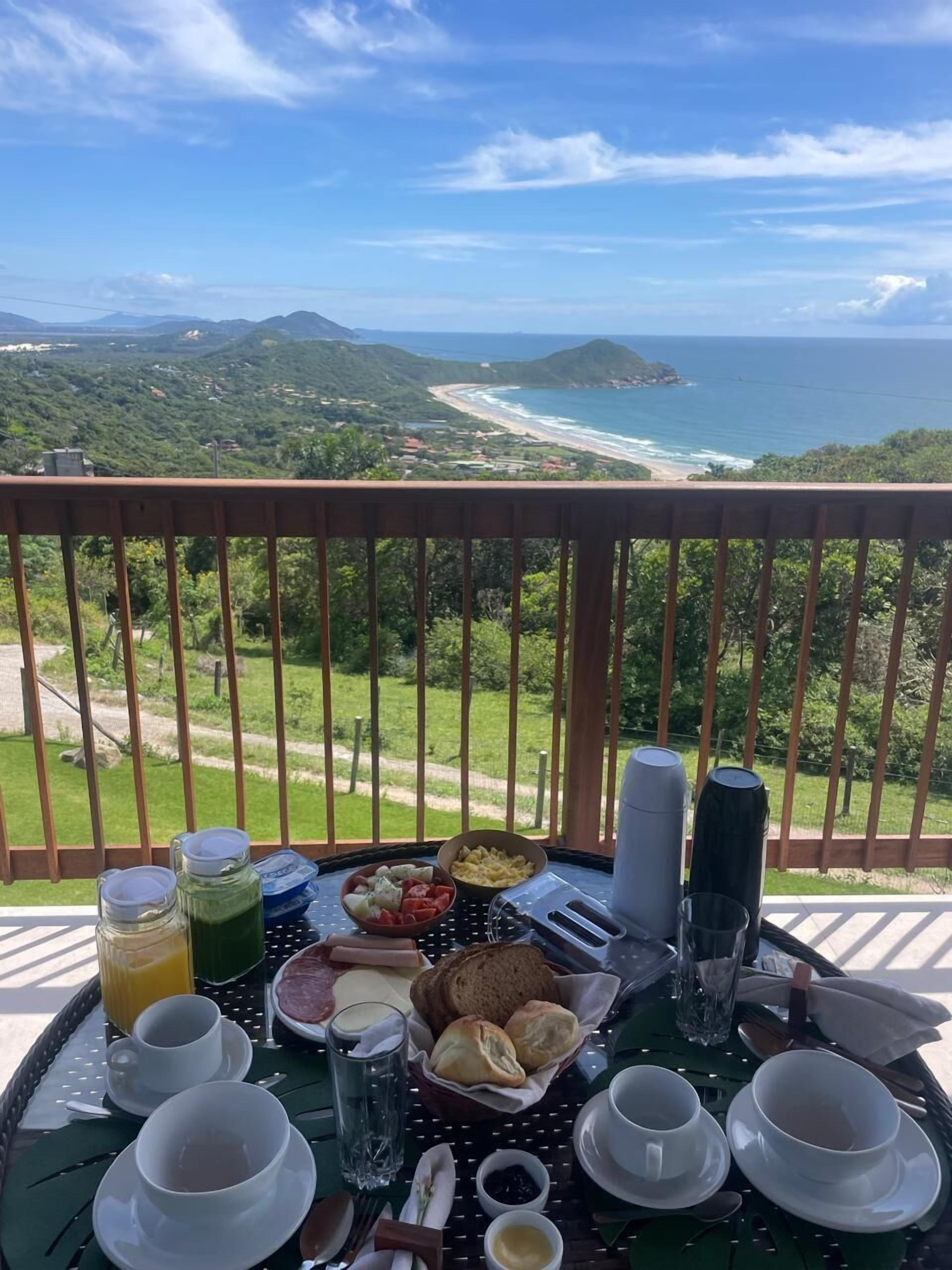 Una mesa de desayuno con diversas comidas y bebidas, con vistas a un paisaje costero con colinas y una playa.