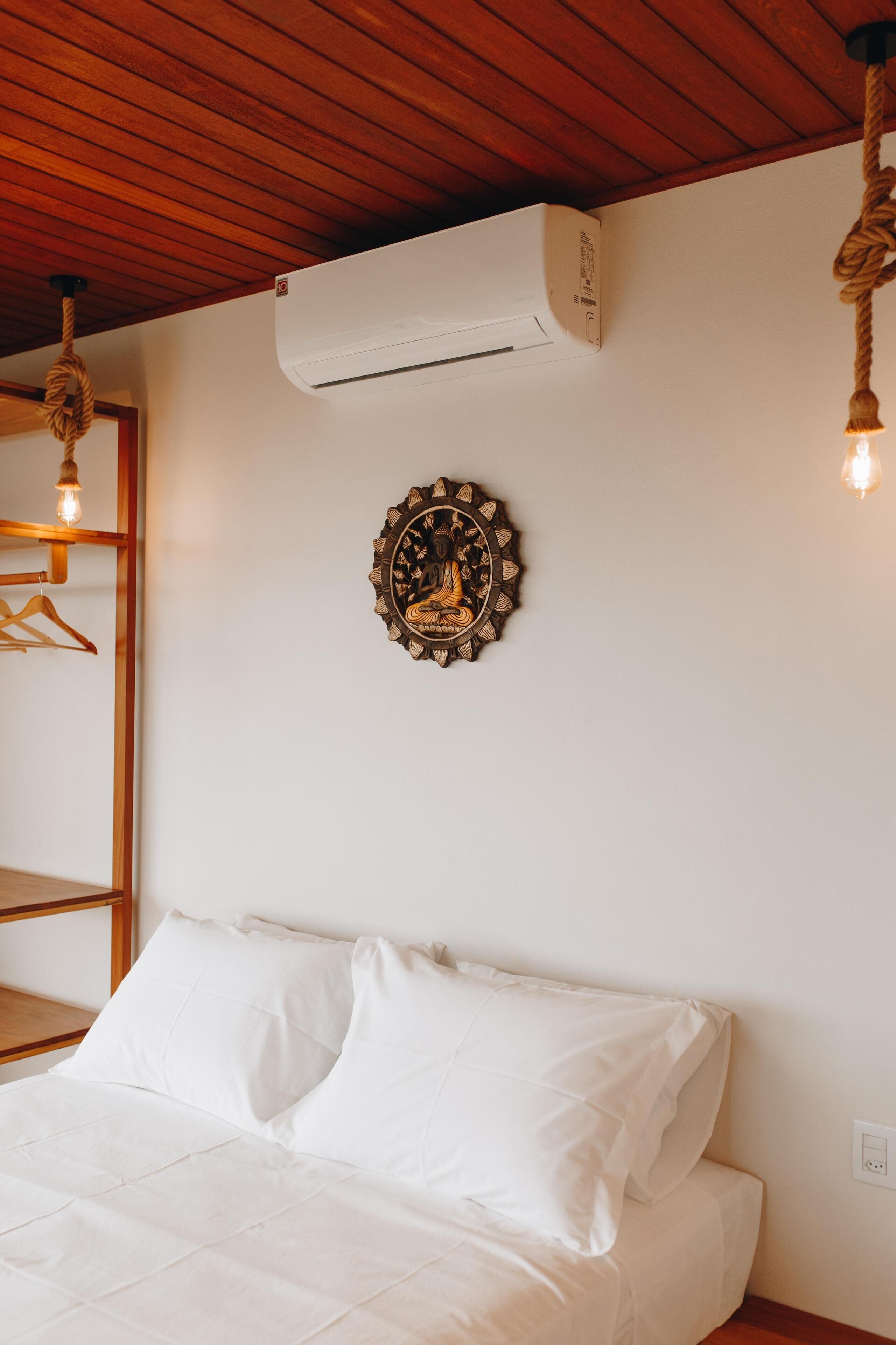 Un dormitorio con techo de madera, una cama con ropa de cama blanca, un aire acondicionado, un adorno en la pared y una bombilla.