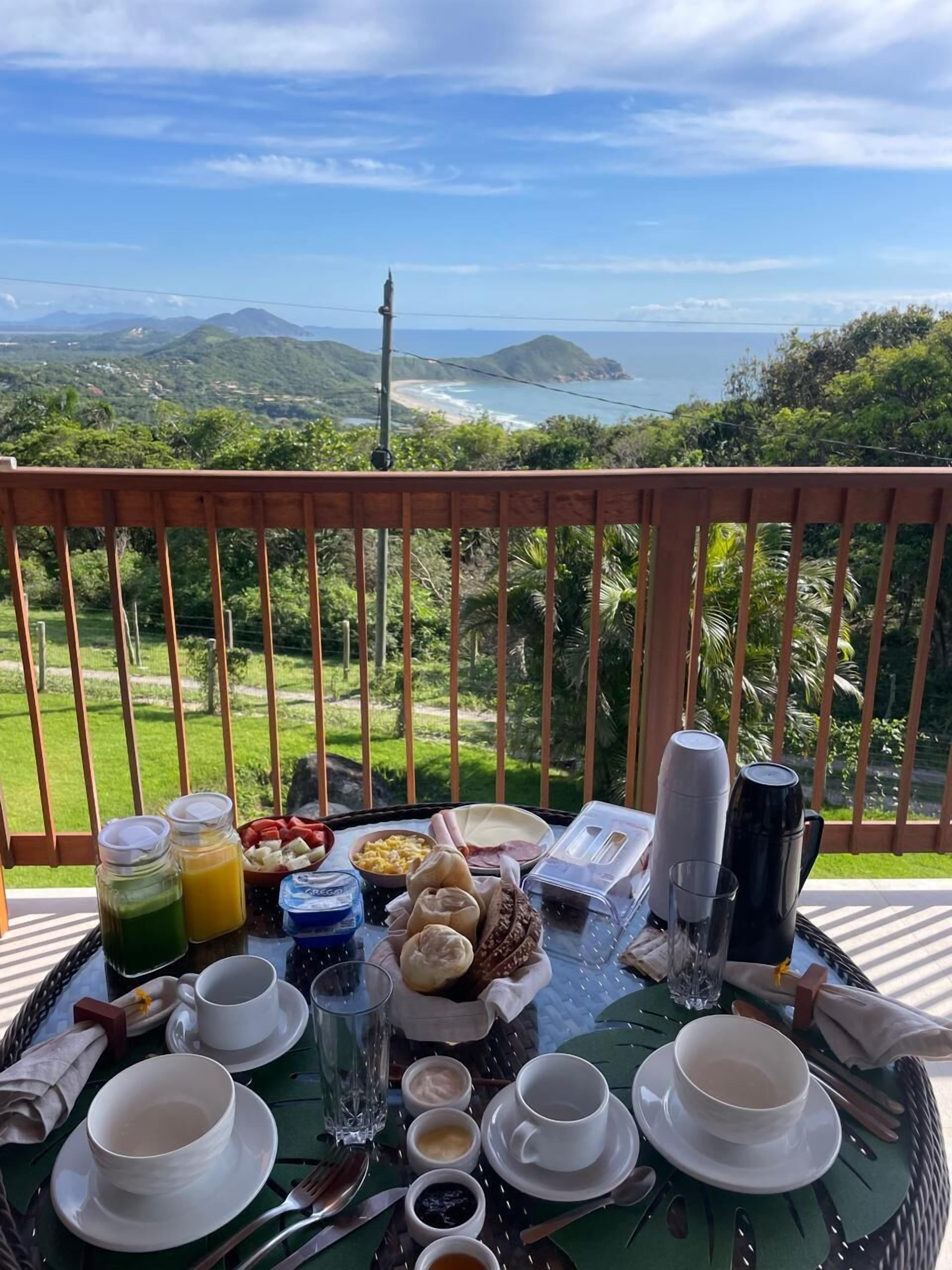 Una mesa de desayuno con diversos alimentos y bebidas, en un balcón con vistas panorámicas al océano y las colinas.
