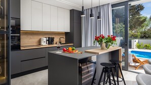 Une cuisine moderne dotée d’un îlot central, d’un four encastré et d’une machine à café. Un coin repas offre une vue sur la piscine, visible à travers les portes vitrées.