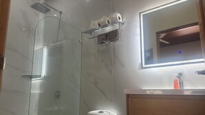 A bathroom with a glass-enclosed shower, a toilet, a vanity with a mirror, and a soap dispenser.