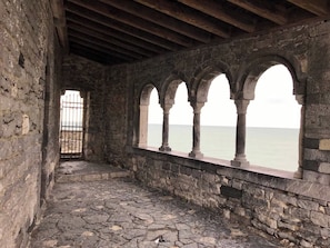 Un couloir en pierre, orné d’arches, mène à une entrée fermée par un portail et offre une vue sur la mer.