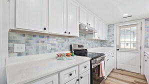 A modern kitchen with white cabinets, a tiled backsplash, stainless steel appliances, and a window with blinds. - 2nd Row, Beach Cottage, 3BR/2BA!  (Murrells Inlet)