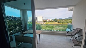 Un balcón con puerta de cristal, una mesa, dos sillas y vista a un campo verde y edificios a lo lejos.
