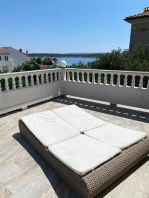 Un balcone con un divano in vimini e cuscini, con vista su uno specchio d'acqua e vegetazione.