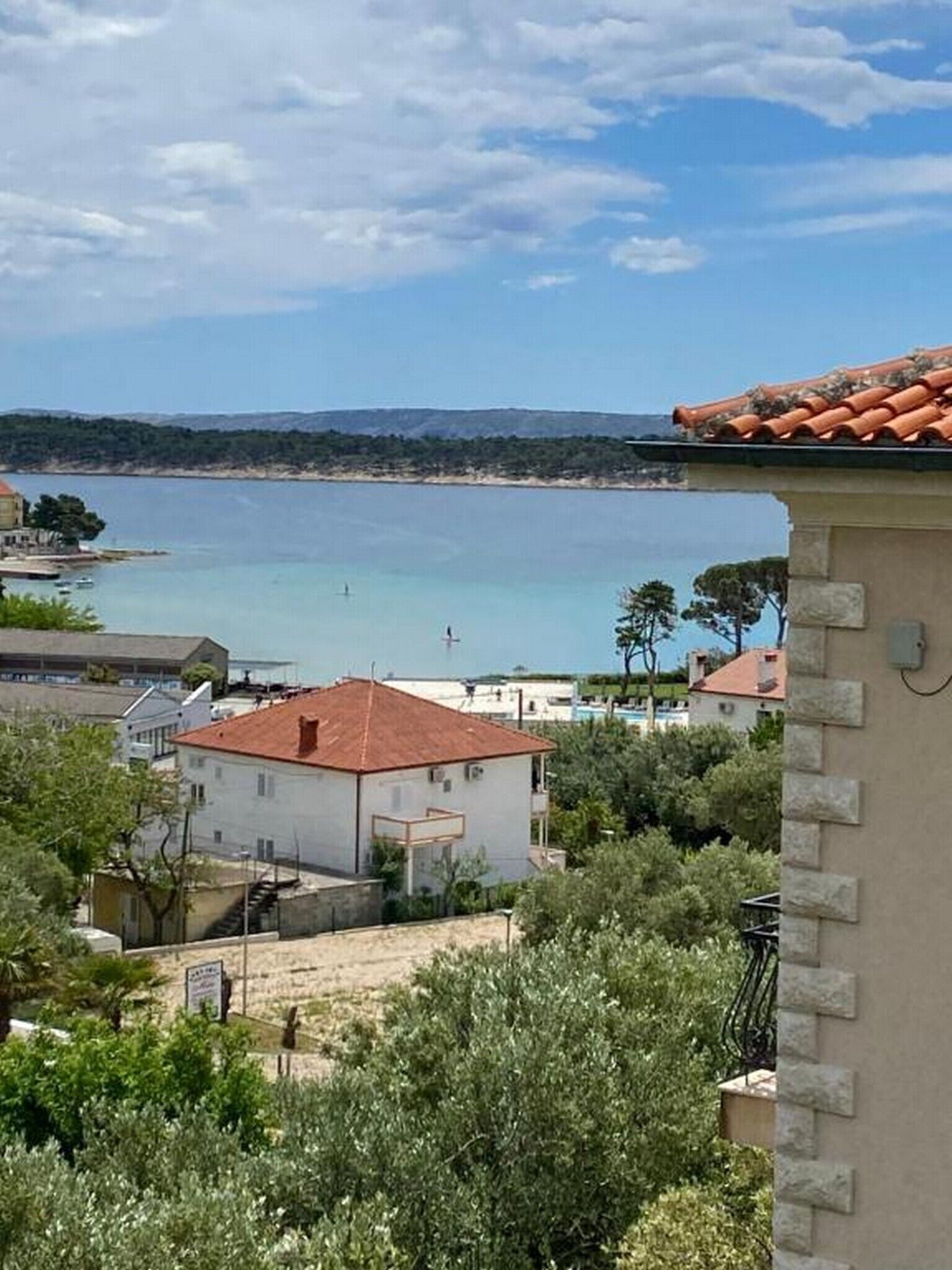 A coastal view with a building featuring a red-tiled roof, surrounded by greenery and overlooking a calm sea.
