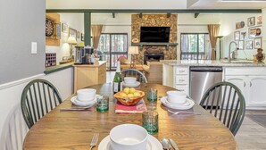Un comedor con una mesa de madera dispuesta para cuatro personas, rodeada de sillas. Se observa una chimenea de piedra con un televisor encima al fondo.
