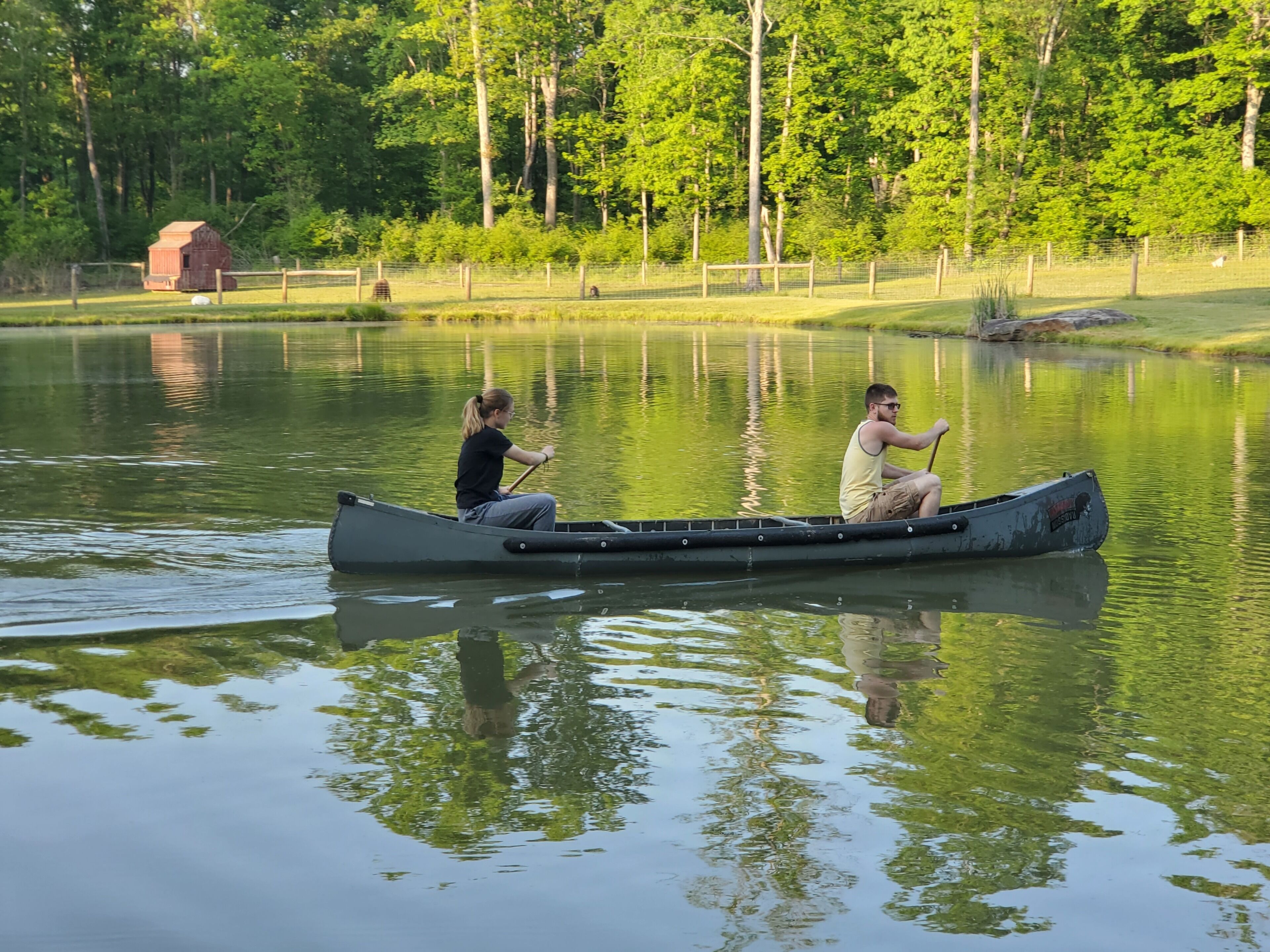 Dos personas en una canoa sobre un lago tranquilo, rodeado de árboles y una estructura de madera al fondo.