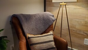 A brown leather armchair with a knitted throw and a striped pillow, positioned next to a small round table with a patterned top. A floor lamp with a white shade stands nearby, and a wooden wall panel is visible in the background.
