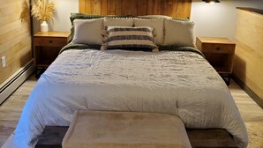 A bedroom with a wooden headboard, a large bed with white bedding, two bedside tables with lamps, and a painting above the bed.