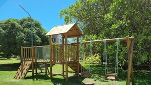 A wooden playground structure with slides and swings, surrounded by grass and trees. - ILOA: Conforto, Natureza e Lazer (Barra de São Miguel)