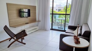 A modern living room with a flat-screen TV mounted on a wooden panel, a sofa, a small round table with a model ship, and a balcony with a chair. - ILOA: Conforto, Natureza e Lazer (Barra de São Miguel)