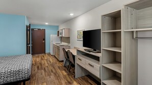 Une chambre de taille modeste comprenant un lit, un bureau, une chaise, un téléviseur, une armoire et une petite kitchenette.