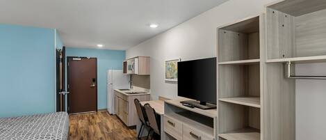 Une chambre de taille modeste comprenant un lit, un bureau, une chaise, un téléviseur, une armoire et une petite kitchenette.