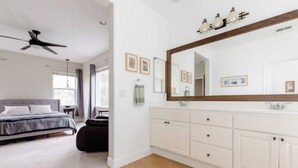 A bedroom with a bed, a ceiling fan, and a large mirror above a white vanity with cabinets. - Luxury Beach Home with Hot Tub
 (San Diego)