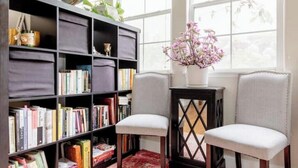 A room with a bookshelf filled with books, two upholstered chairs, a small table with a vase of flowers, and a window with a view of greenery. - Luxury Beach Home with Hot Tub
 (San Diego)
