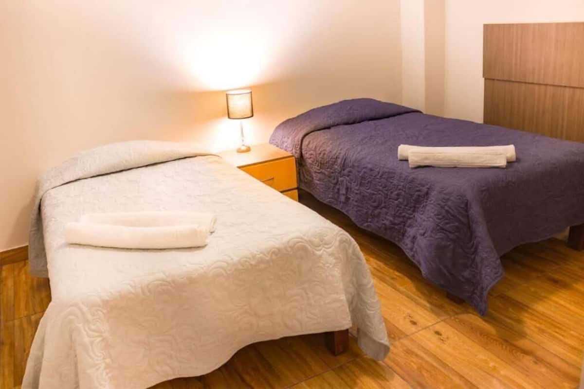 Dos camas individuales con cabeceras, una mesita de noche con lámpara y un armario de madera en una habitación con suelo de madera.