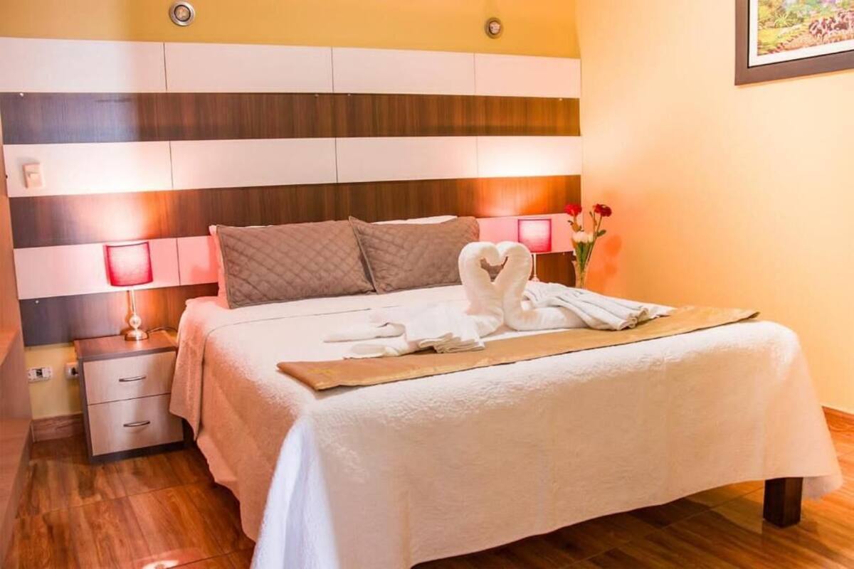 Una cama bien hecha con una toalla dispuesta en forma de corazón, una lámpara de noche y una mesita de noche con cajones.