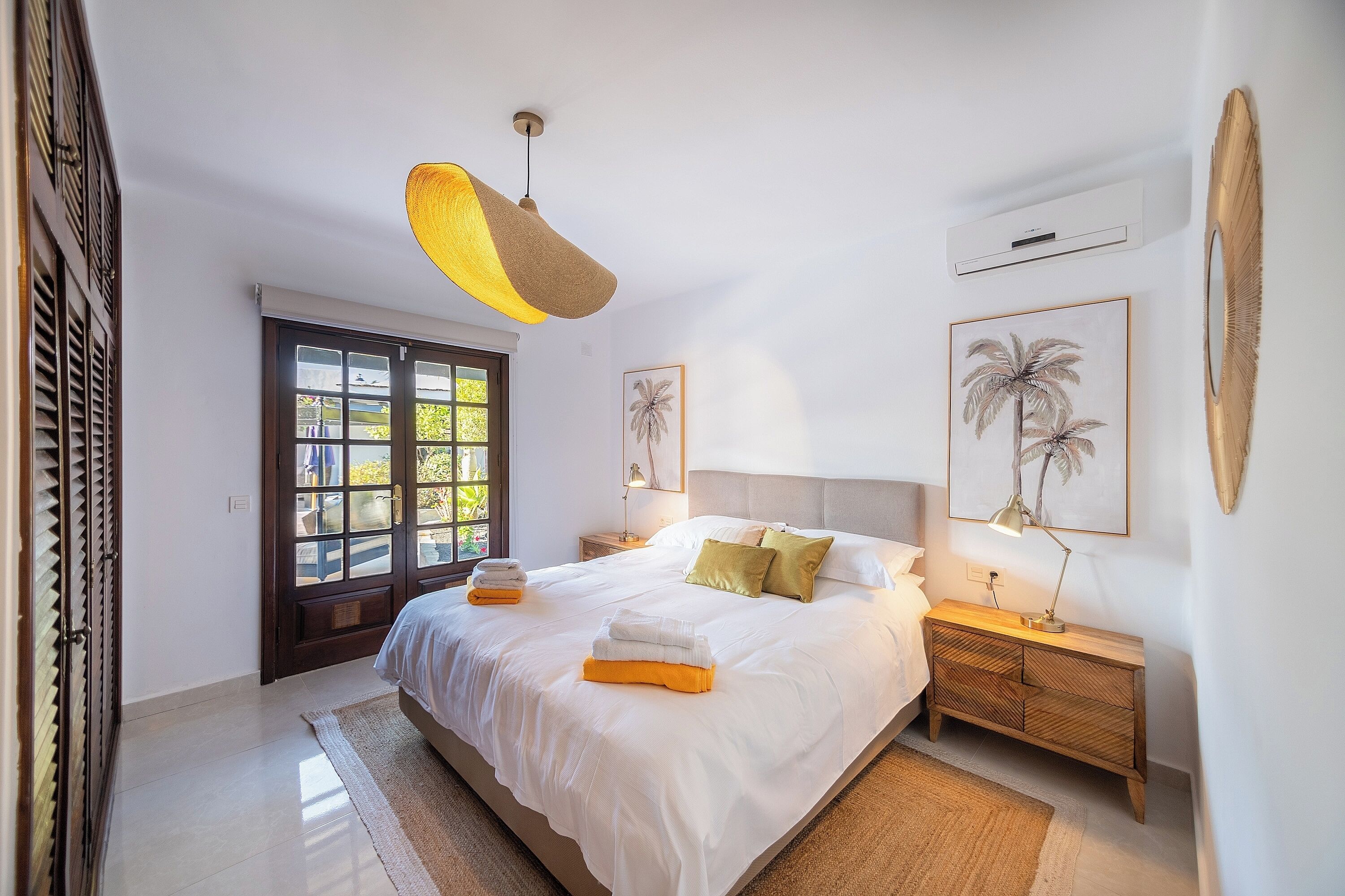 A spacious bedroom with a large bed, a wooden nightstand, a hanging light fixture, and a view through a glass door.