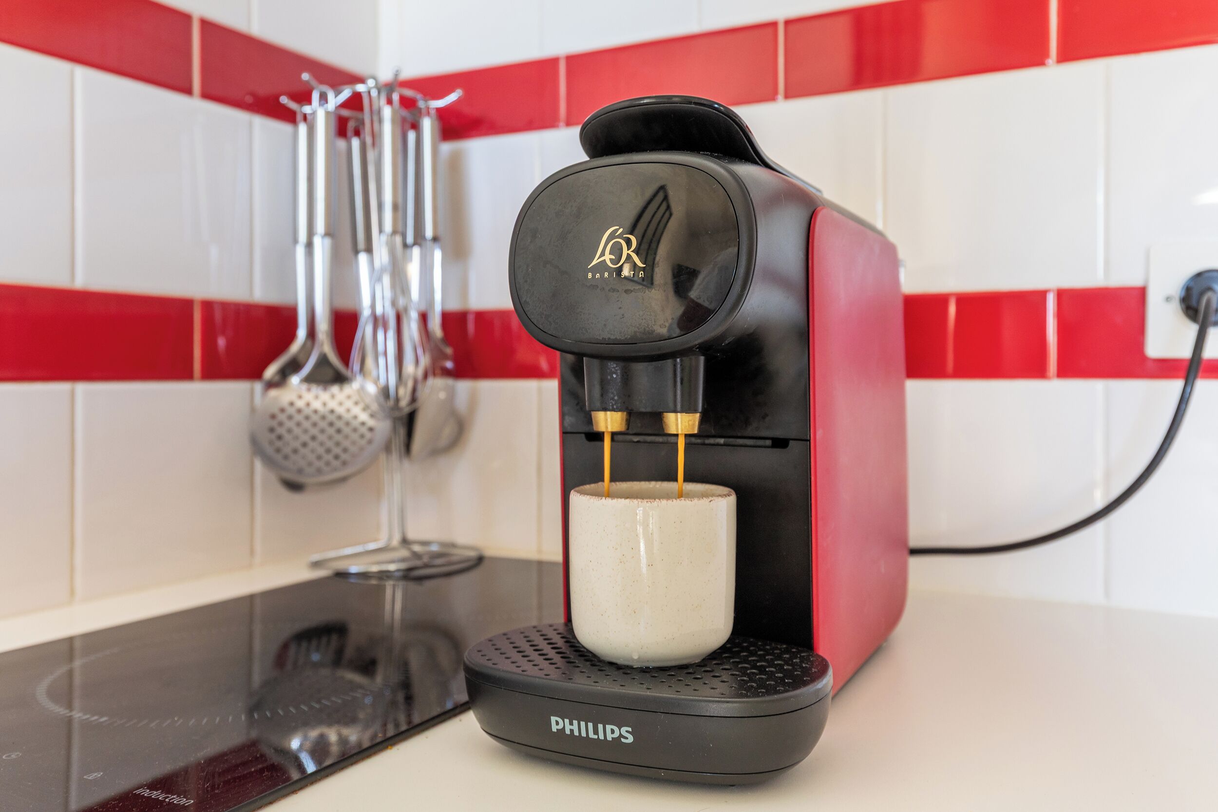 Une machine à café dotée d’un porte-tasse, des ustensiles de cuisine et un pare-éclaboussures carrelé de carreaux rouges et blancs.